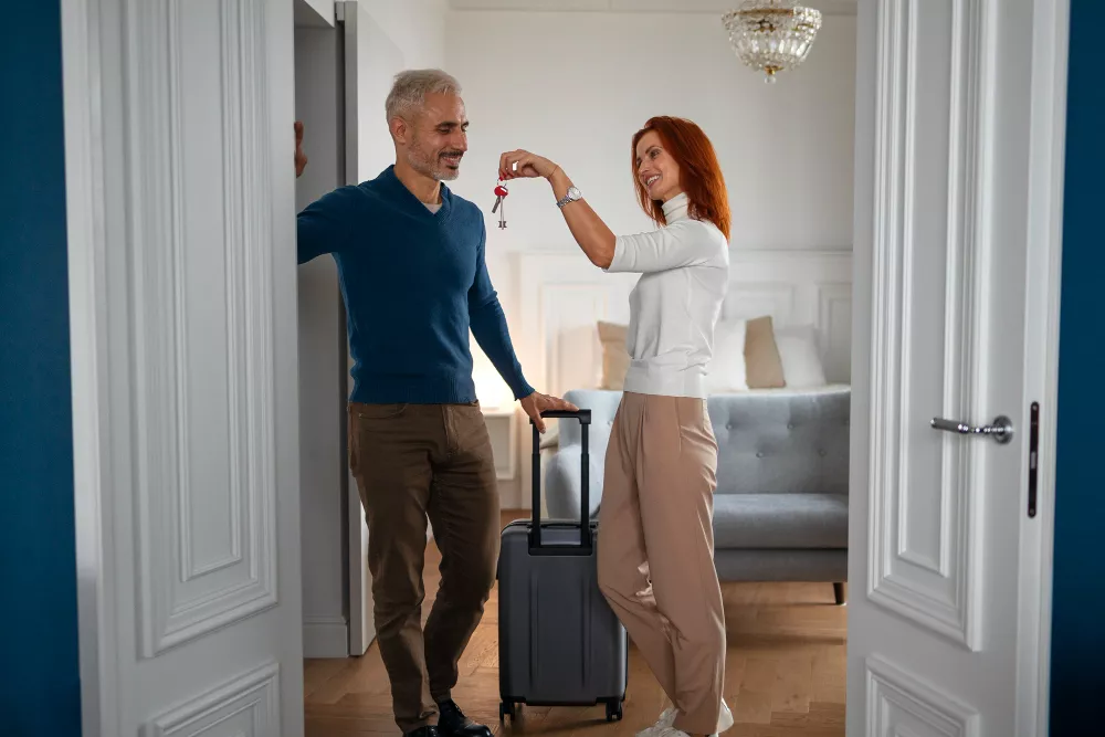Happy couple arriving at a vacation rental, holding keys and luggage in a bright, modern living room.