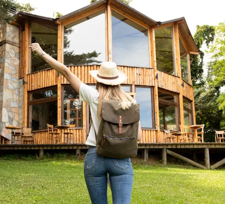 Traveler raising hand with keys in front of a wooden cabin vacation rental surrounded by trees.
