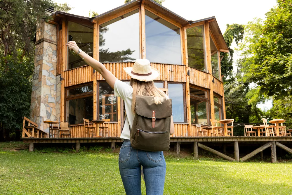 Traveler raising hand with keys in front of a wooden cabin vacation rental surrounded by trees.
